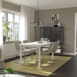 Modern dining room with white STRANDTORP extendable table, grey chairs, glass cabinet, and green rug. Large window for natural light.
