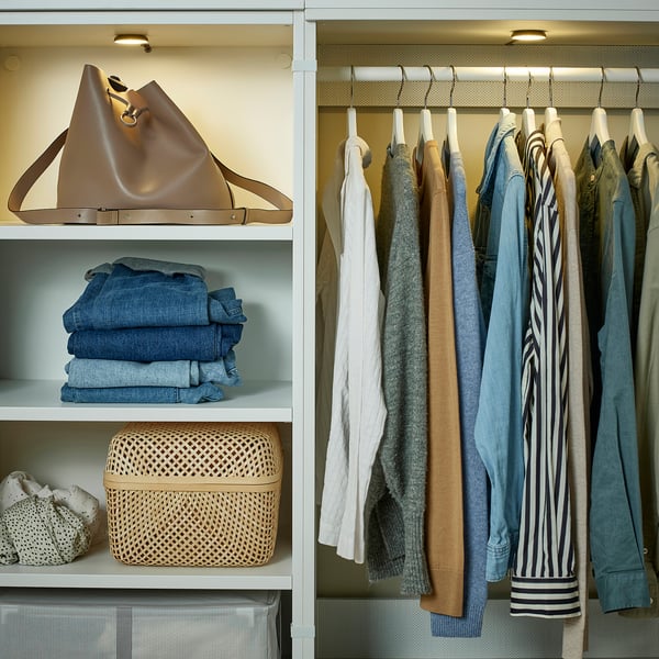 Organized closet with lit shelves. Clothes hang neatly on right, folded jeans stack below large beige handbag, additional wicker bin on low shelf.