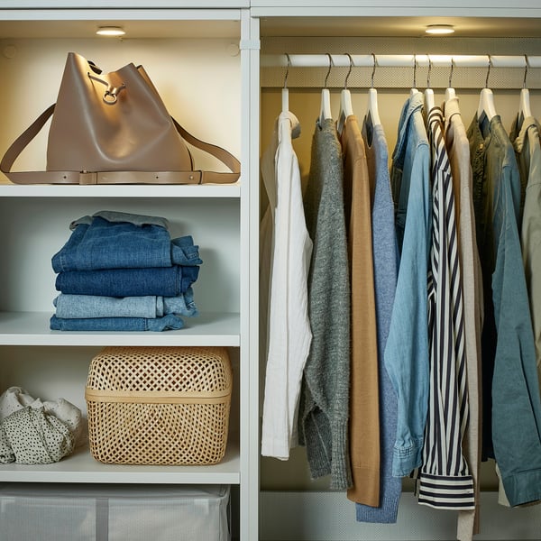 Organized closet with clothes and accessories. Spotlight illuminates shelves featuring a large handbag, folded jeans, and a woven basket.