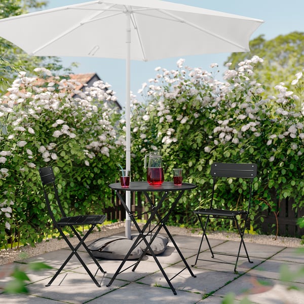 Outdoor patio setup with black foldable table and chairs, white umbrella, red drinks, gray cushions, surrounded by blooming white flowers.
