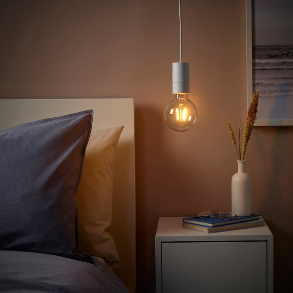 Cozy bedroom with SUNNEBY pendant lamp over bedside table, warm light, books, dried flowers, and blue-yellow pillows.