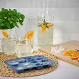 Ice cube tray with frozen shapes sits on woven mat next to glasses of citrus drink.