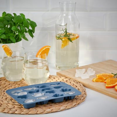 Ice cube tray with frozen shapes sits on woven mat next to glasses of citrus drink.