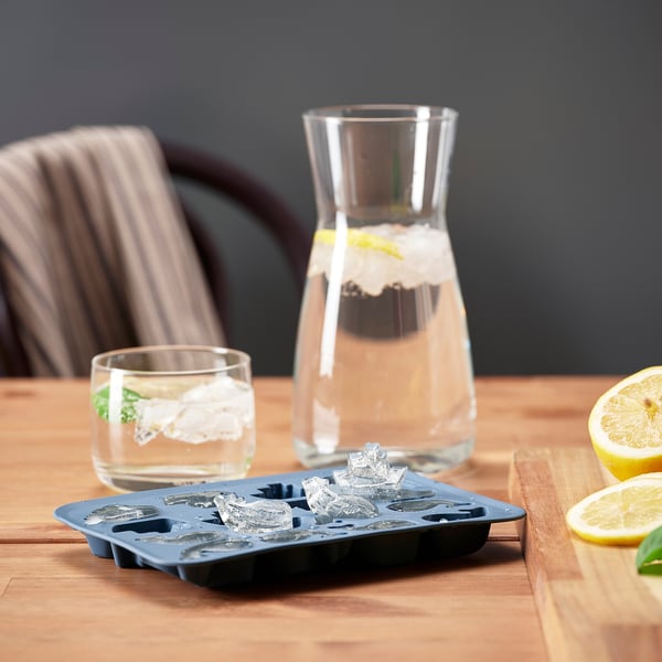 Ice cube tray with playful shapes on wooden table, next to a glass of water and a pitcher, perfect for summer drinks.