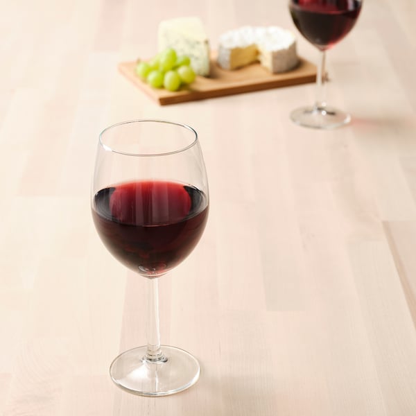 Two wine glasses with red wine on a wooden table. One glass near a cheese board with grapes and cheeses.