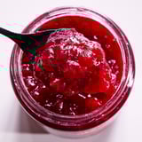 Open jar of bright red lingonberry jam, spoonful lifted, glass container.