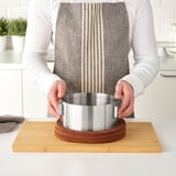 Person uses adjustable metal baking ring to shape cake on wooden board in kitchen.