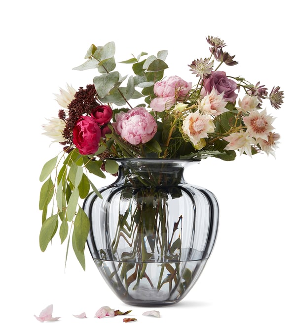 Glass vase with flower bouquet, petals on white surface. Alternative: vase with floral bouquet on white table, petals scattered.