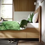 A modern wooden bed with soft-close drawers beneath, featuring a green striped comforter and tropical leaf pillows, set against a wooden panel wall.