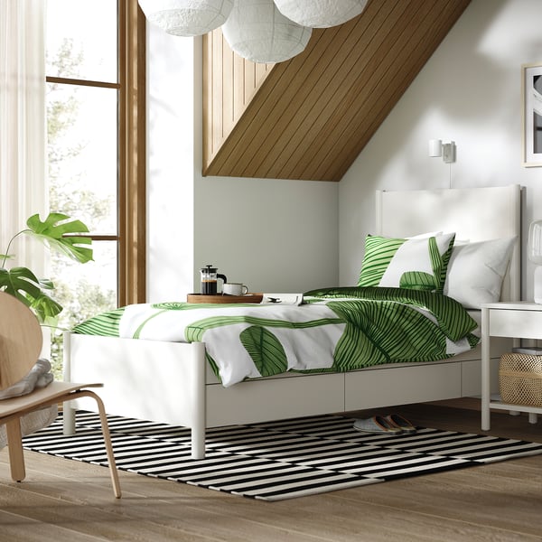 Modern minimalist bedroom with white TONSTAD bed, green leaf bedding, black & white striped rug, wooden floor, large window, and hanging lights.