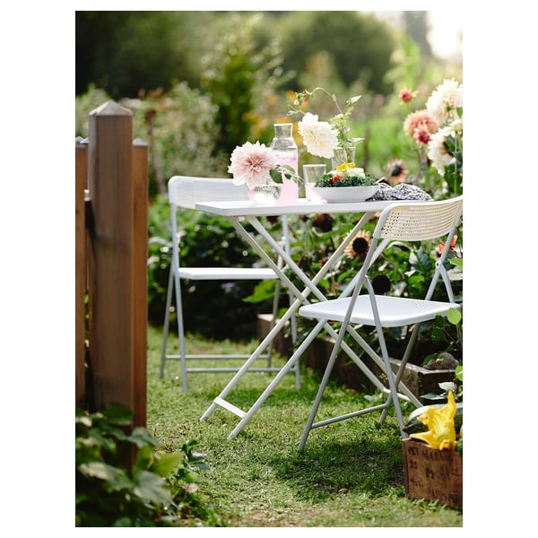 Image description: white metal outdoor table and chairs set in a garden with colorful flowers and wooden fence.
