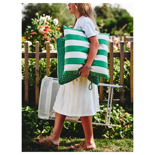 A woman carries green and white striped outdoor cushions near a white folding chair on a grassy area with a wooden fence and plants.