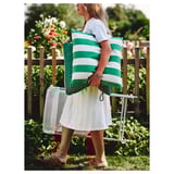 A woman carries green and white striped outdoor cushions near a white folding chair on a grassy area with a wooden fence and plants.