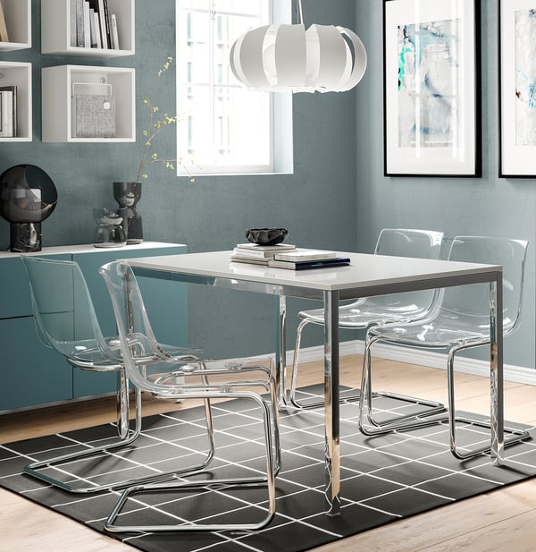 Modern dining set with clear chairs, glossy white table, chrome legs, under cabinet lighting, blue wall, and abstract art.