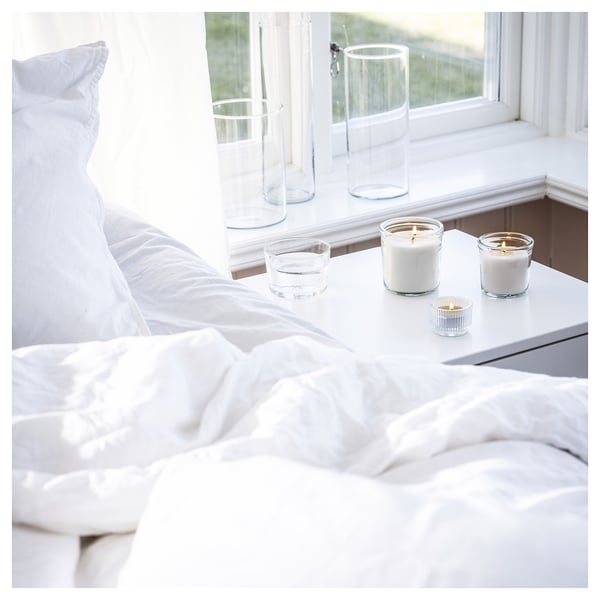 Cozy bedroom scene with white bedding, four lit candles on nightstand near window, sunlight streaming in.