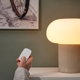 Hand holding white remote near lit beige lamp on white table, green wall with bike photo.