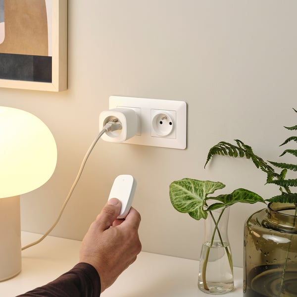 A hand holds a remote controlling a lamp plugged into a white socket, beside a potted plant.