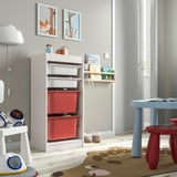 A TROFAST storage unit with white frame and red boxes, placed in a kids playroom. It holds toys and books, and is next to a small table and stools.