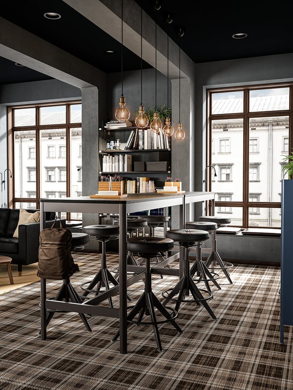Modern office with high black tables and chairs, light gray walls, large windows, hanging lights, shelves with books, and leather bag.