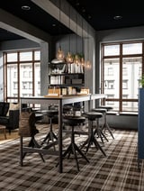 Modern office with high black tables and chairs, light gray walls, large windows, hanging lights, shelves with books, and leather bag.