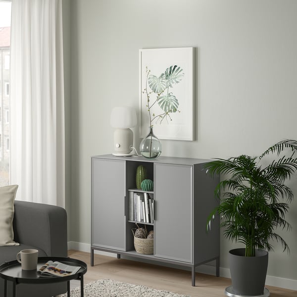 TULLSTORP metal sideboard in living room, gray with shelves and beveled doors, holding books and decor, beside a green plant.