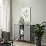 TULLSTORP metal sideboard in living room, gray with shelves and beveled doors, holding books and decor, beside a green plant.