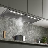 Modern kitchen with stainless hood emitting steam, integrated above marble backsplash, lighted and functional.