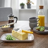 Breakfast setup with cheese slicer, cheese, cucumber, coffee, oj, and fruit on table.