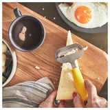 Person holds yellow UPPFYLLD cheese slicer, slicing cheese near breakfast items.
