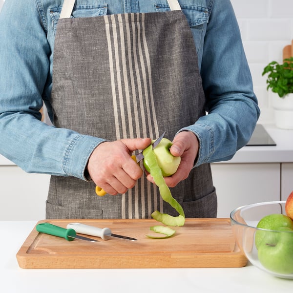 Person peeling green apple with small paring knife from UPPFYLLD set on wooden cutting board. Bowl of peeled apples nearby.