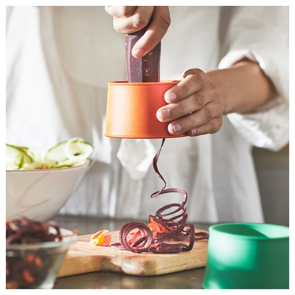 Person using orange UPPFYLLD vegetable slicer to cut red vegetable into spiral strips on wooden board.