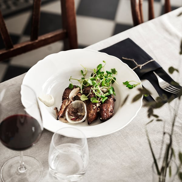 A white plate with grilled food and greens on a table with wine and cutlery.