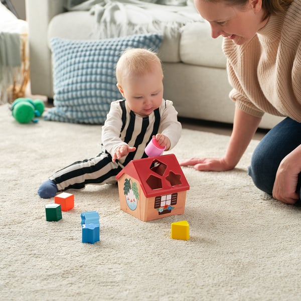 Baby playing with shape sorter toy, guided by adult. Child holds pink block near wooden house-shaped toy with colored blocks around.