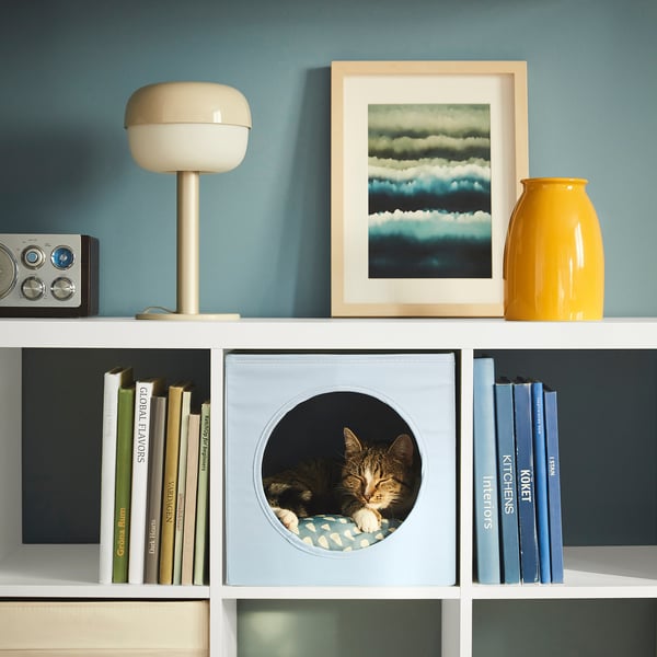 A white shelf with cat house containing a cat, books, a lamp, a radio, and a vase.