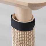 A cylindrical cat scratching mat made of woven material wraps around a table leg, secured with black straps for cats to stretch and sharpen claws.