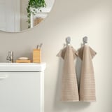 Beige VÅGSJÖN towels hang on hooks beside a white vanity, mirror, and toothbrush holder.