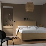 Modern minimalist bedroom with large wooden bed under woven pendant light, white bedding, wooden dresser with mirror, dark chair, and beige rug.