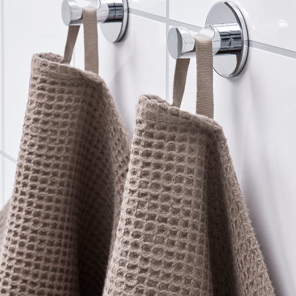 Two beige towels with textured weave hang neatly on silver hooks, showcasing their absorbent, quick-drying material and fringe edging.