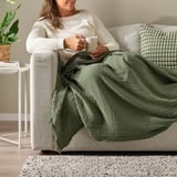 A woman relaxes on a beige sofa, holding a cup of coffee. Shes wrapped in a green VALLKRASSING throw blanket.