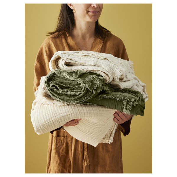 A woman holding a stack of soft, crinkled VALLKRASSING throws in white and green. The throws have raw edges and are made of 100% cotton.