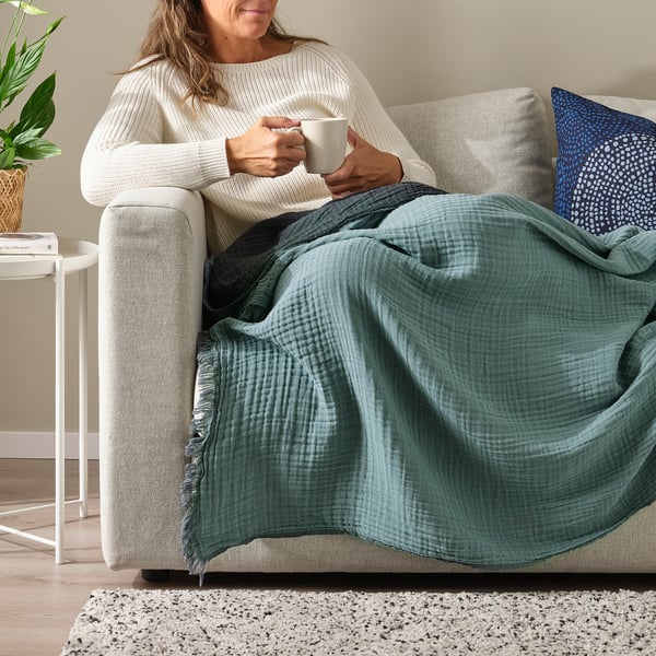 Woman relaxes on couch sipping coffee under soft, crinkled green VALLKRASSING throw.