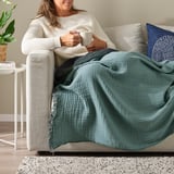 Woman relaxes on couch sipping coffee under soft, crinkled green VALLKRASSING throw.