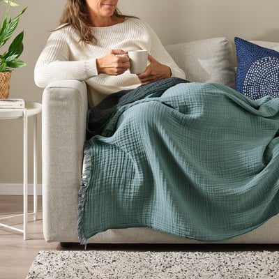 Woman relaxes on couch sipping coffee under soft, crinkled green VALLKRASSING throw.
