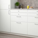 Modern white kitchen with VALLSTENA drawers, stainless handles, wooden floor, sandwich, juice, and herb plant.