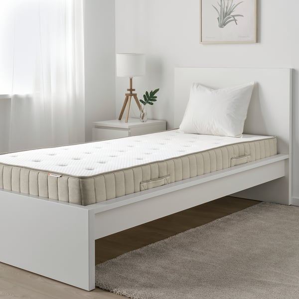 Minimalist bedroom with white mattress, small nightstand, lamp, and plant.