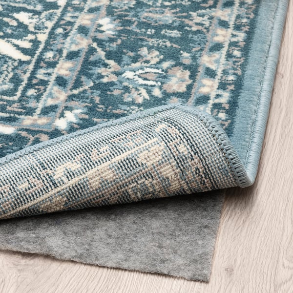A blue patterned rug laid on a wooden floor, partially folded to show its anti-slip underlay, demonstrating its functional grip and design.