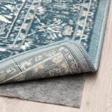 A blue patterned rug laid on a wooden floor, partially folded to show its anti-slip underlay, demonstrating its functional grip and design.