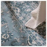 A blue VEDBÄK rug with white floral patterns, made of synthetic fibers, placed on a textured surface.