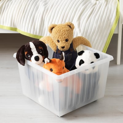 Clear plastic storage bin with wheels, holding various stuffed animals, sits on a wooden floor beside a bed.