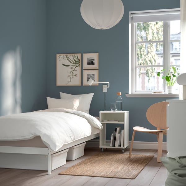Minimalist bedroom with white VEVELSTAD bed, blue walls, natural rug, wooden chair, side table with books, lamp, and plants near window.